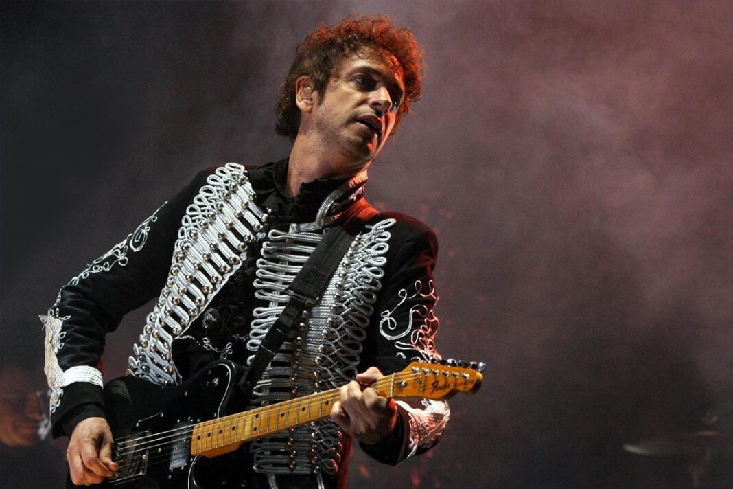 ></noscript> Gustavo Cerati en vivo con traje blanco durante la gira Fuerza Natural, tocando la guitarra bajo luces tenues.