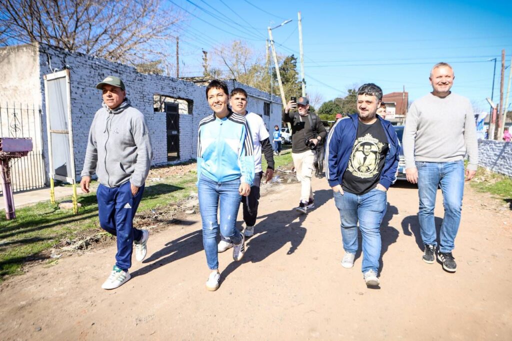 Mayra Mendoza y Facundo Tignanelli caminando por una calle de tierra en el conurbano bonaerense junto a vecinos y militantes.