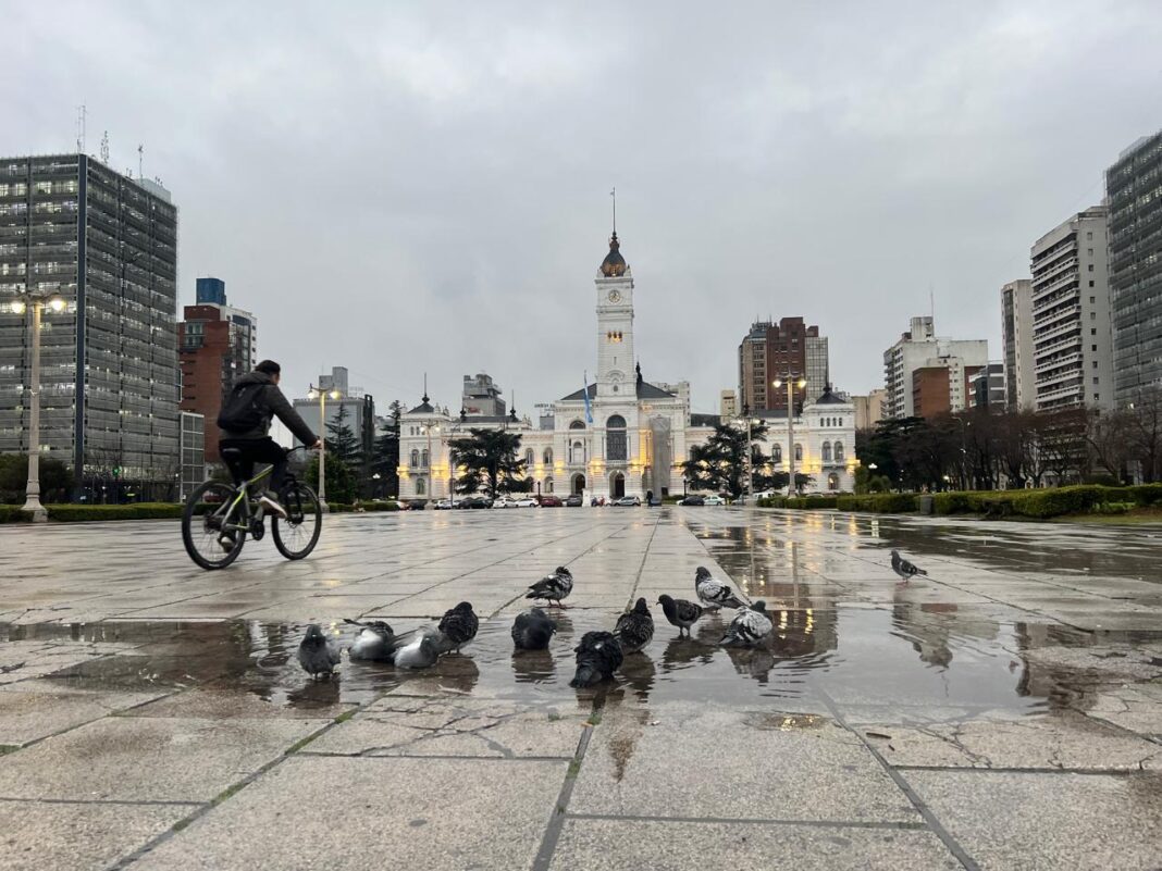 Ciclista atraviesa Plaza Moreno en La Plata en una mañana nublada y húmeda, con la Municipalidad de fondo