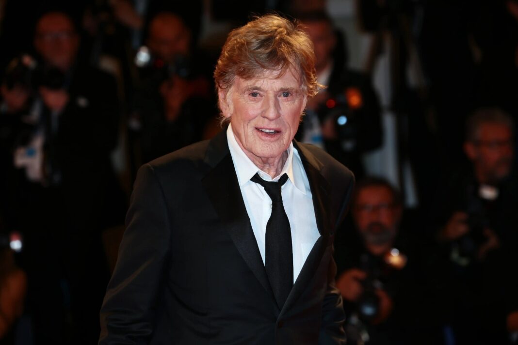 Robert Redford con traje negro en una alfombra roja, rodeado de fotógrafos.