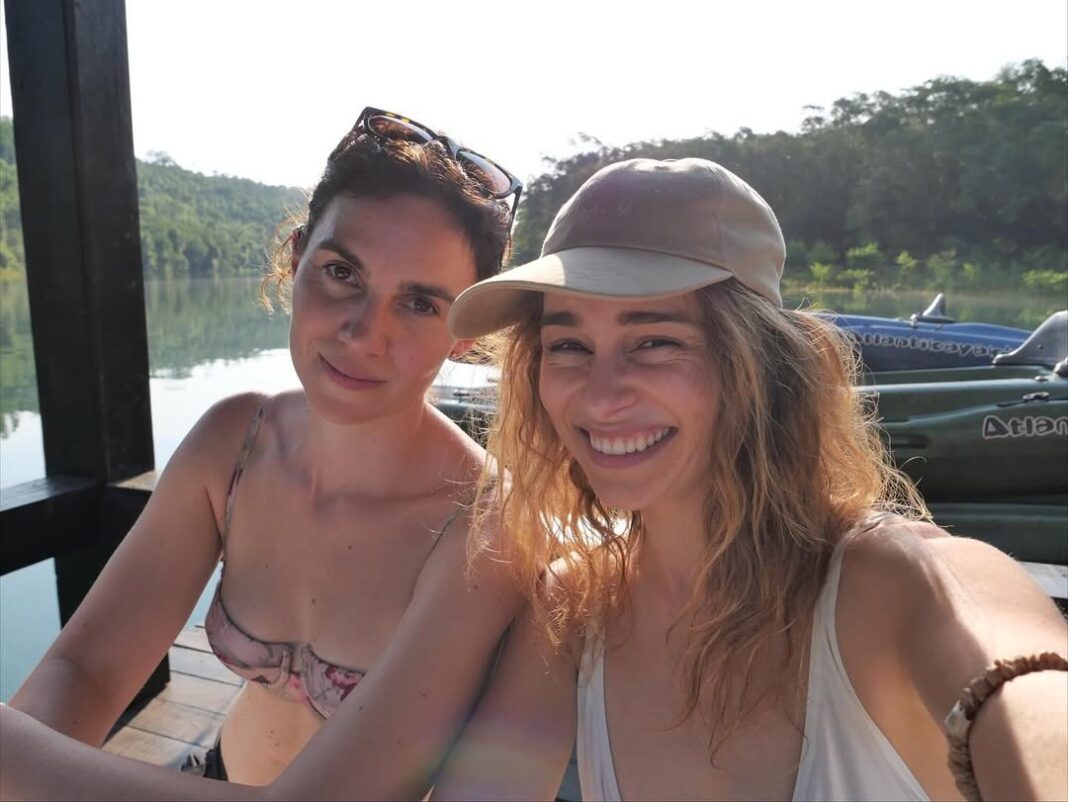 Emilia Clarke sonriendo en selfie con amiga en Iguazú, Argentina, con kayaks y naturaleza de fondo.