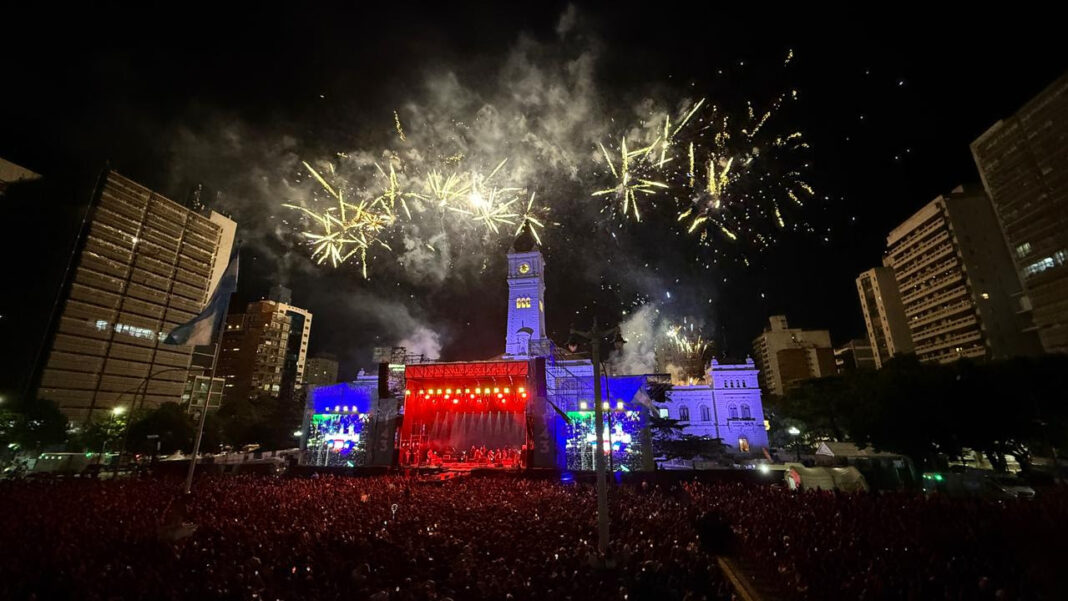 Multitud masiva de más de 100 mil personas disfrutando de un concierto nocturno en Plaza Moreno de La Plata, con fuegos artificiales iluminando el cielo sobre el edificio histórico con torre del reloj.