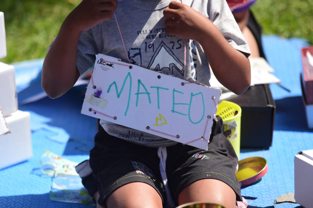 Niño pequeño sentado al aire libre sobre una alfombra azul, sosteniendo un cartel hecho a mano que dice 