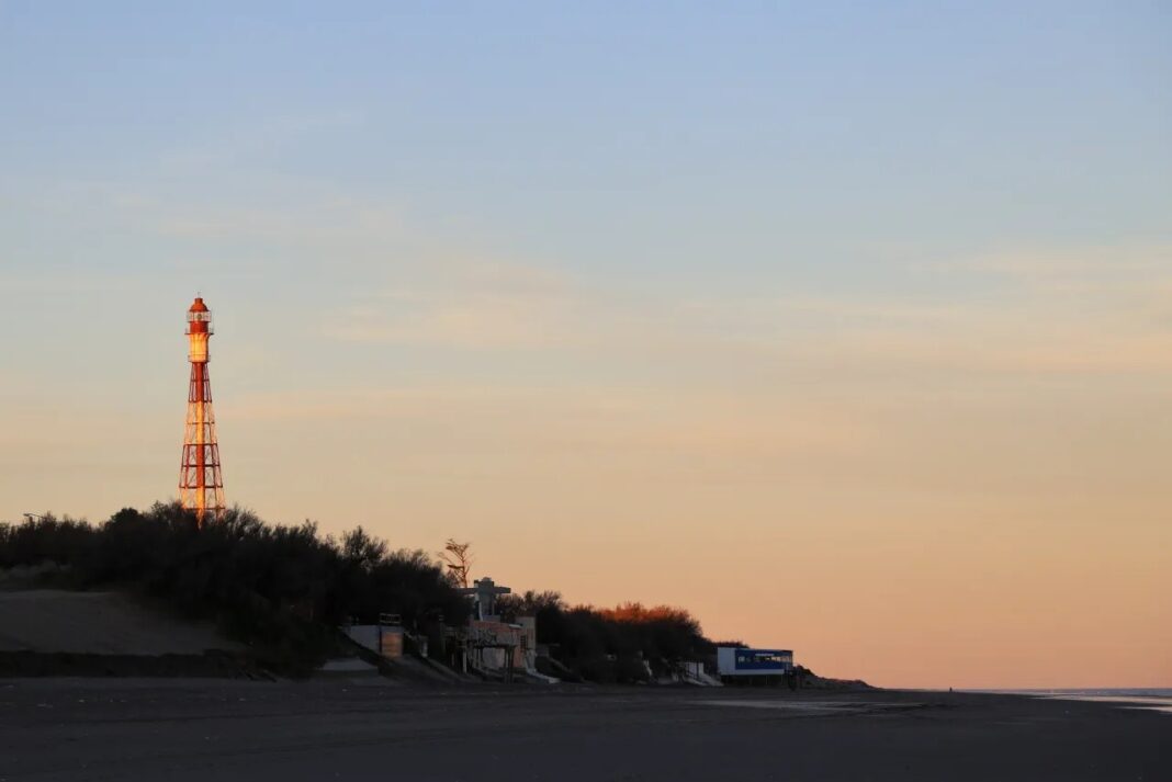 Impresionante faro de celosía rojo y blanco (Faro Recalada) en Monte Hermoso, Costa Atlántica, destacándose sobre el follaje oscuro durante un atardecer/amanecer.