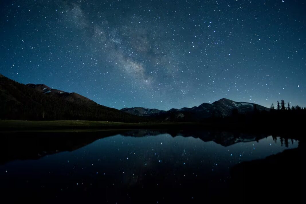 Impresionante fotografía de larga exposición de la Vía Láctea arqueándose sobre montañas boscosas reflejadas en un lago tranquilo, ilustrando la intensidad de la Luna Nueva en Escorpio.