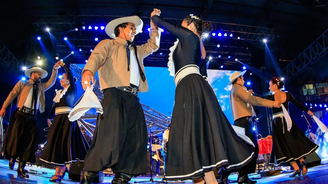 Grupo de bailarines con vestimenta tradicional de gaucho (faldas negras y sombreros) realizando una danza folklórica en el escenario del Pre Cosquín iluminado con luces azules.