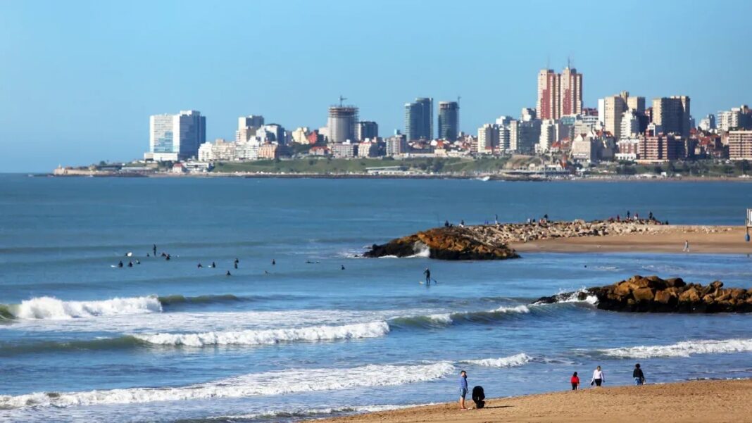 Día soleado en la Costa Atlántica de Buenos Aires con surfistas en el mar, ideal para el último fin de semana largo de 2025.