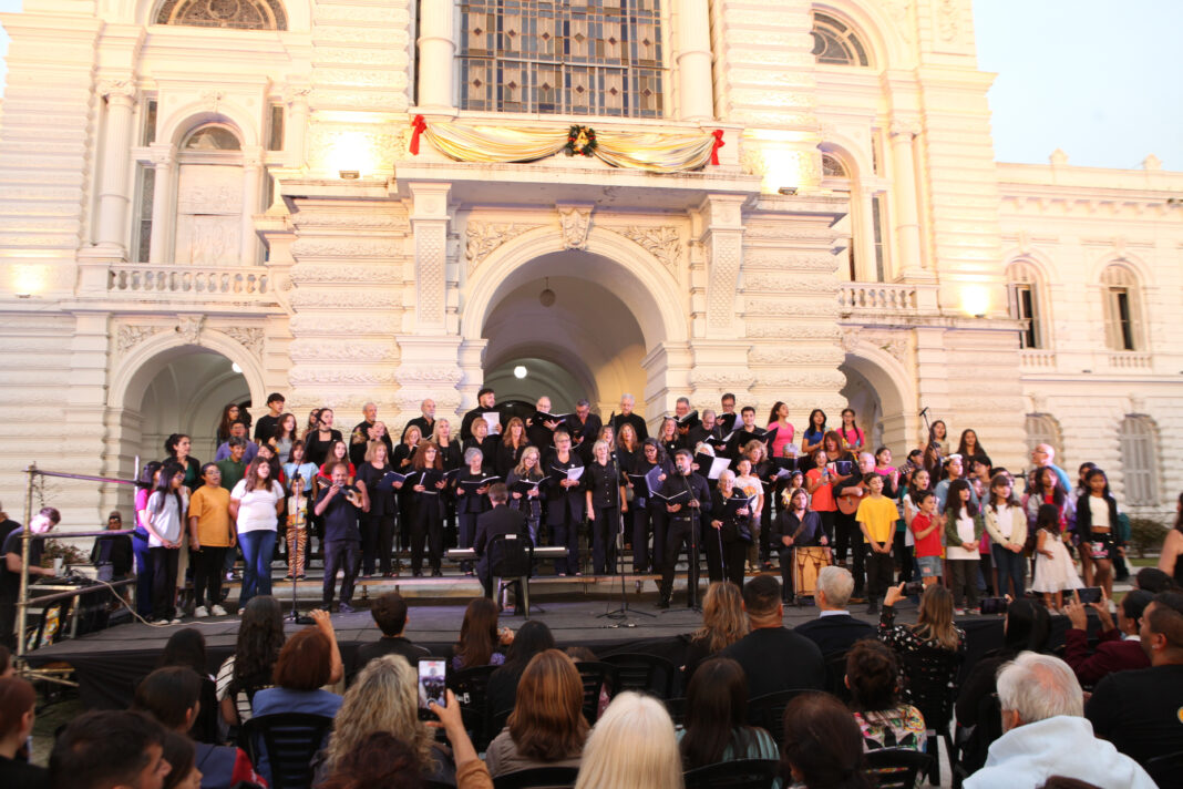 Gran coro de adultos y niños actuando al aire libre frente a un edificio municipal clásico e iluminado durante las celebraciones navideñas.