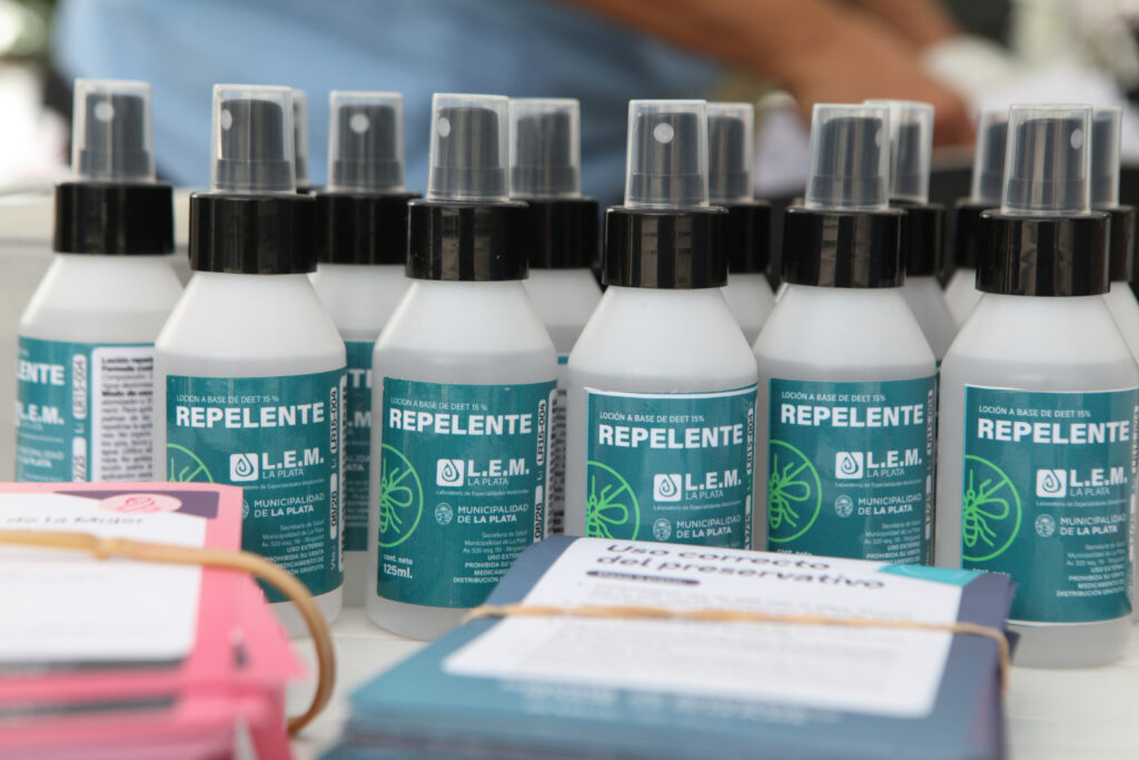 Múltiples envases de repelente en spray plástico blanco con etiqueta "REPELENTE" (15% DEET) de la Municipalidad de La Plata, junto a folletos informativos.